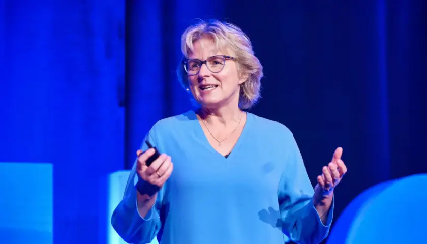 Melanie Wolfers, eine inspirierende Rednerin, hält einen Vortrag vor einem blauen Hintergrund. Sie trägt eine blaue Bluse und eine Halskette.
