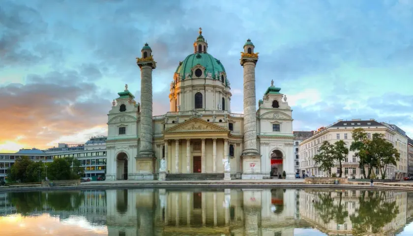 Die Karlskirche Wien spiegelt sich im Wasser. Majestätische Säulen flankieren die Kuppel. Ein dramatischer Himmel über der barocken Pracht.