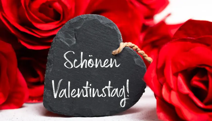 Herzförmiges Schild mit der Aufschrift "Schönen Valentinstag!" vor roten Rosen. Ein liebevoller Gruß zum heiligen Valentin.