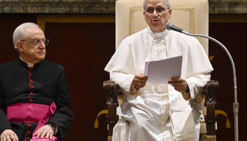 Papst Franziskus spricht in ein Mikrofon, während ein Kardinal daneben sitzt.