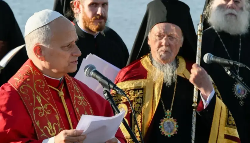 Zwei Geistliche, einer im roten Gewand, der andere mit Bart und Mitra, stehen vor Mikrofonen. Der Mann mit Mitra ist ein Patriarch.