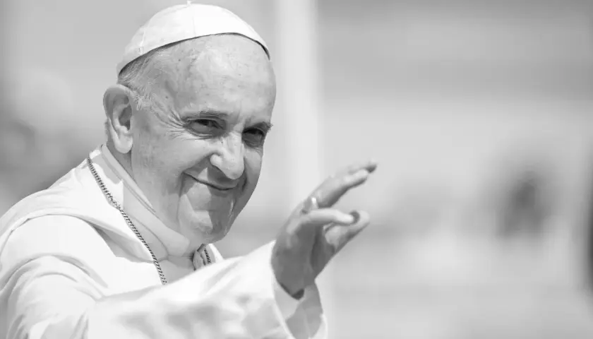 Papst Franziskus winkt lächelnd in die Menge, gekleidet in seine Soutane und mit einer Kette.