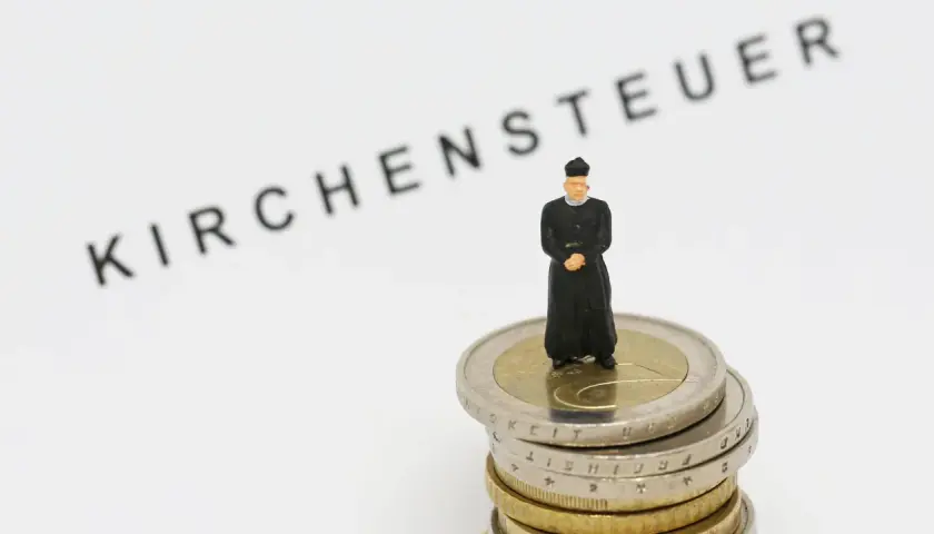 Miniatur-Priester steht auf Münzstapel. Der Schriftzug "Kirchensteuer" deutet auf die Verwendung der Gelder hin.