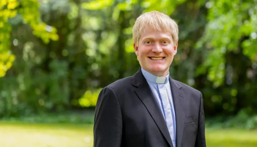 Thomas Frings, ein Geistlicher mit blondem Haar, lächelt freundlich in die Kamera. Er trägt einen schwarzen Anzug mit einem blauen Hemd und einem weißen Kragen.