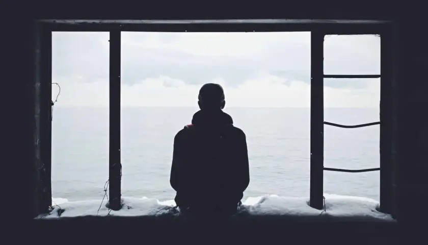 Eine Person blickt aus einem Fenster auf das Meer. Schnee liegt auf dem Sims. Das Bild vermittelt ein Gefühl von Einsamkeit.