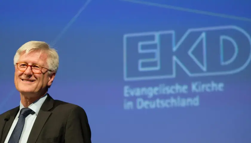 Lächeln des EKD Vorsitzenden vor dem Logo der Evangelischen Kirche in Deutschland.