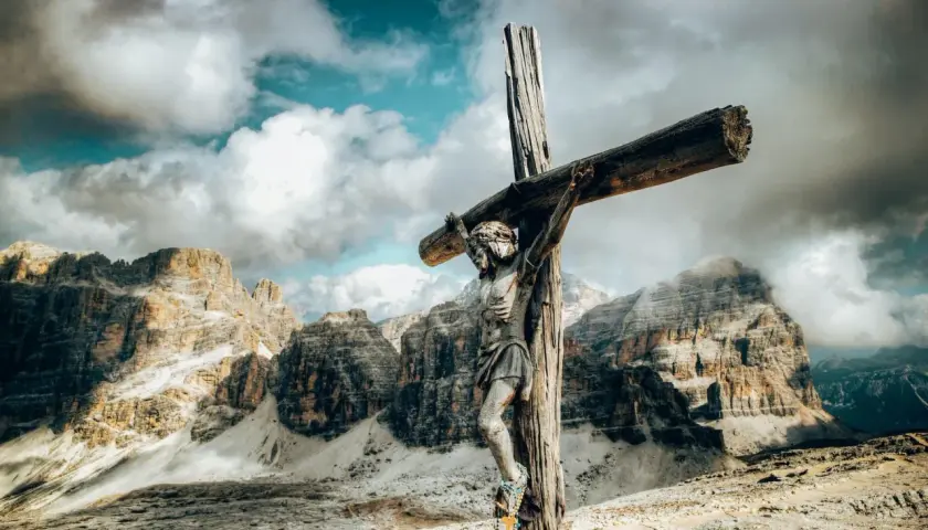 Holzfigur der Jesus-Kreuzigung vor dramatischer Bergkulisse und Wolkenhimmel.
