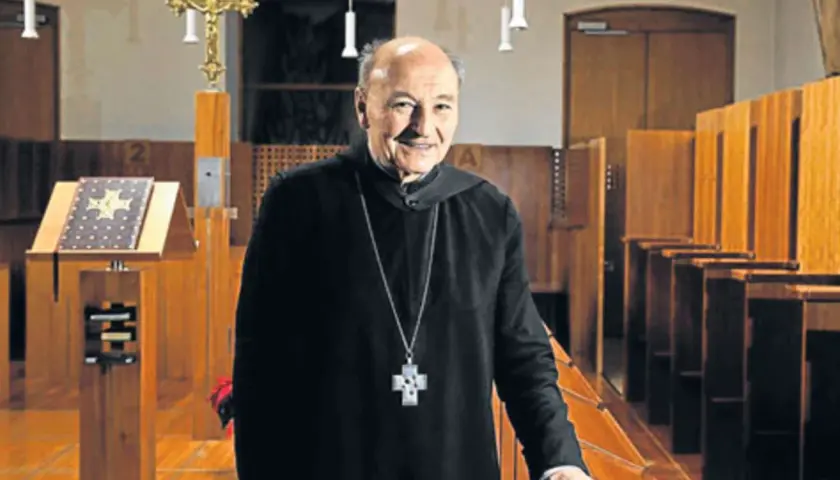 Ein lächelnder Odilo Lechner in schwarzer Soutane mit Kreuzkette steht in einer Kirche vor Beichtstühlen und einem Lesepult.
