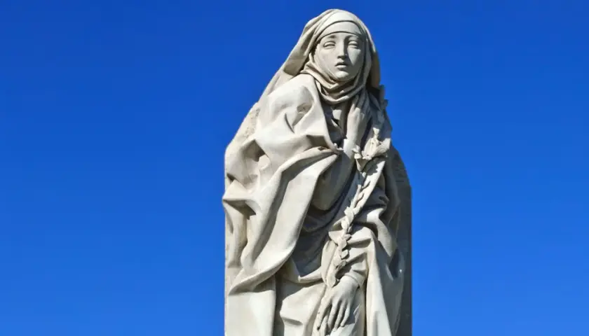 Steinskulptur der Heiligen Katharina von Siena mit Schleier und einem Lorbeerkranz vor blauem Himmel.