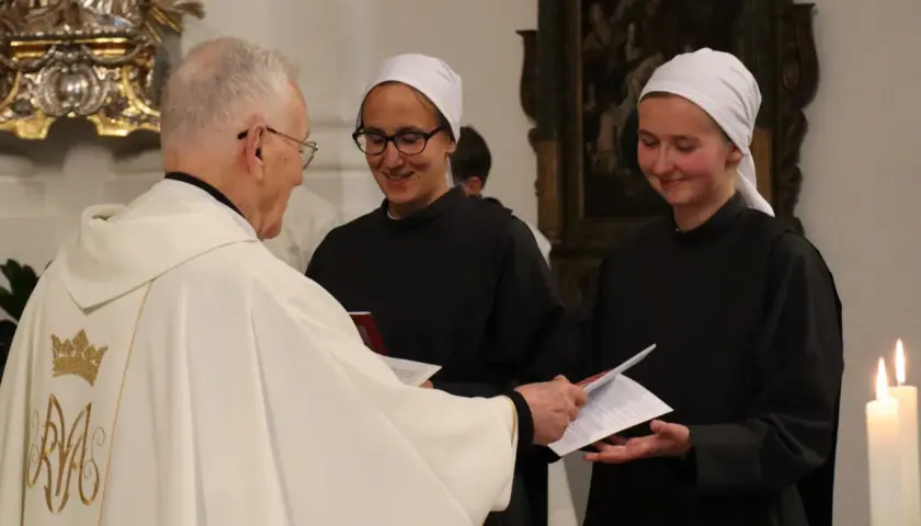 Priester übergibt Dokumente an zwei Dienerinnen vom Heiligen Blut, die lächelnd ihre Aufgaben annehmen.