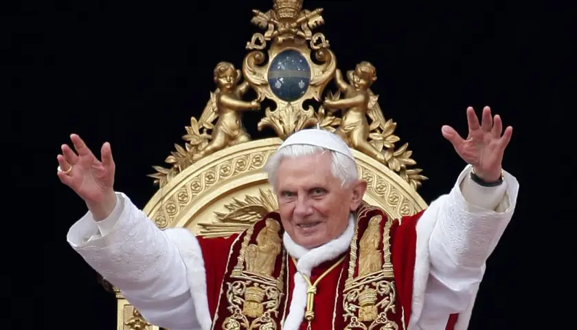 Papst Benedikt XVI. winkt in prächtiger Papst-Kleidung, mit weißer Soutane und rotem Umhang, verziert mit goldenen Stickereien.