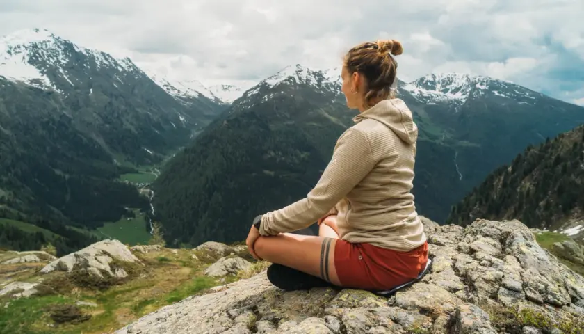 Frau meditiert auf einem Felsen mit Blick auf ein alpines Tal. Sie findet Ruhe und Inspiration für ihre Meditationstexte unterwegs sein.