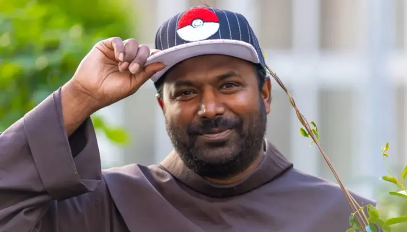 Pater Sandesh Manuel, ein Mann mit Bart, trägt eine Kappe mit Pokémon-Motiv und eine braune Robe. Er lächelt und hebt die Kappe an.