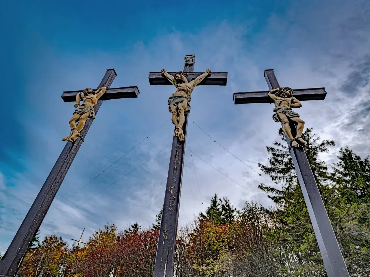Drei Kreuze mit Jesusfiguren, die die Passion Christi darstellen, vor einem dramatischen Himmel und herbstlichem Laub.