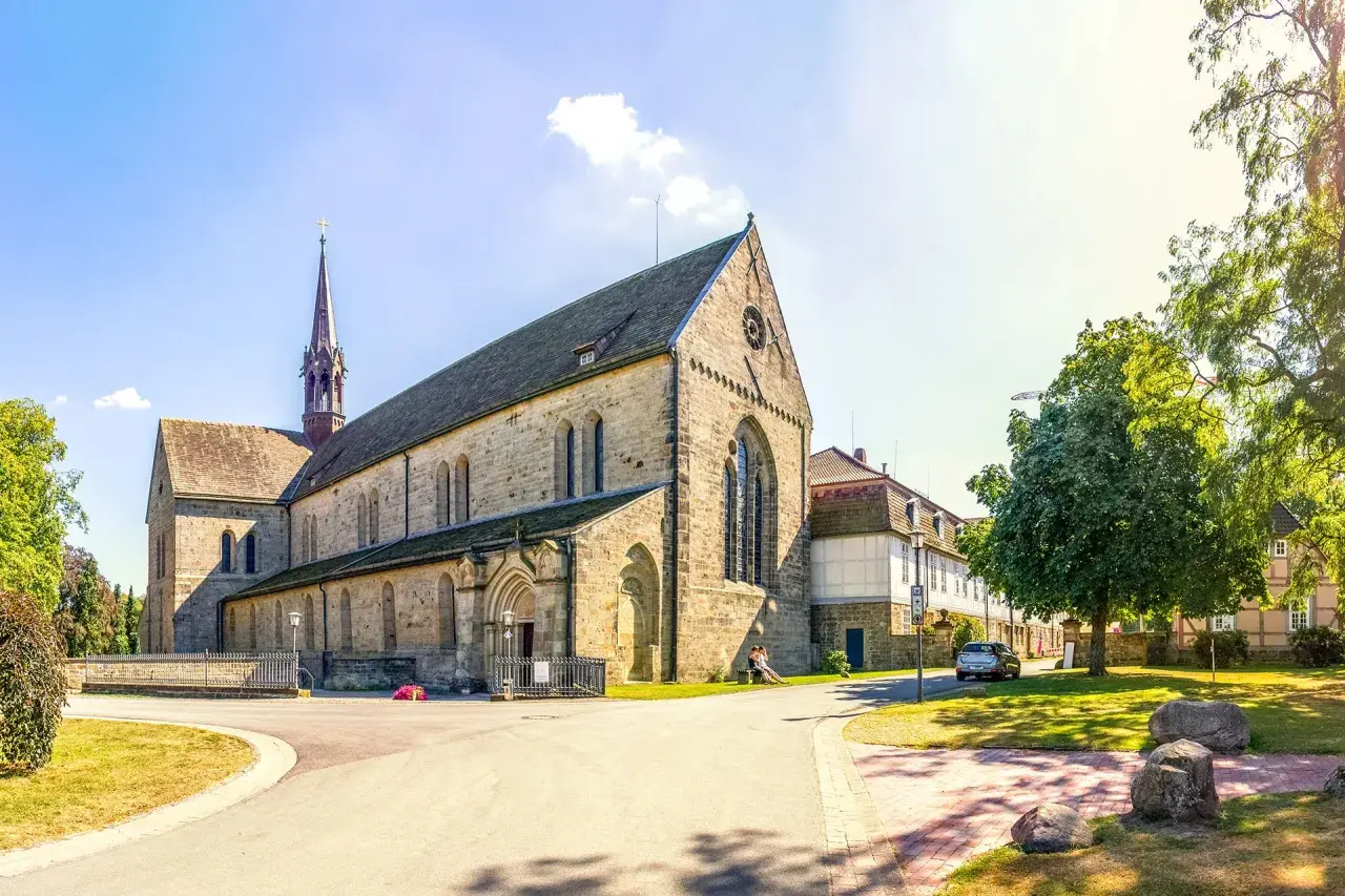Klosterkirche mit Turm, Teil des Jakobsweg Hessen. Ein Paar sitzt auf einer Bank im Schatten eines Baumes.