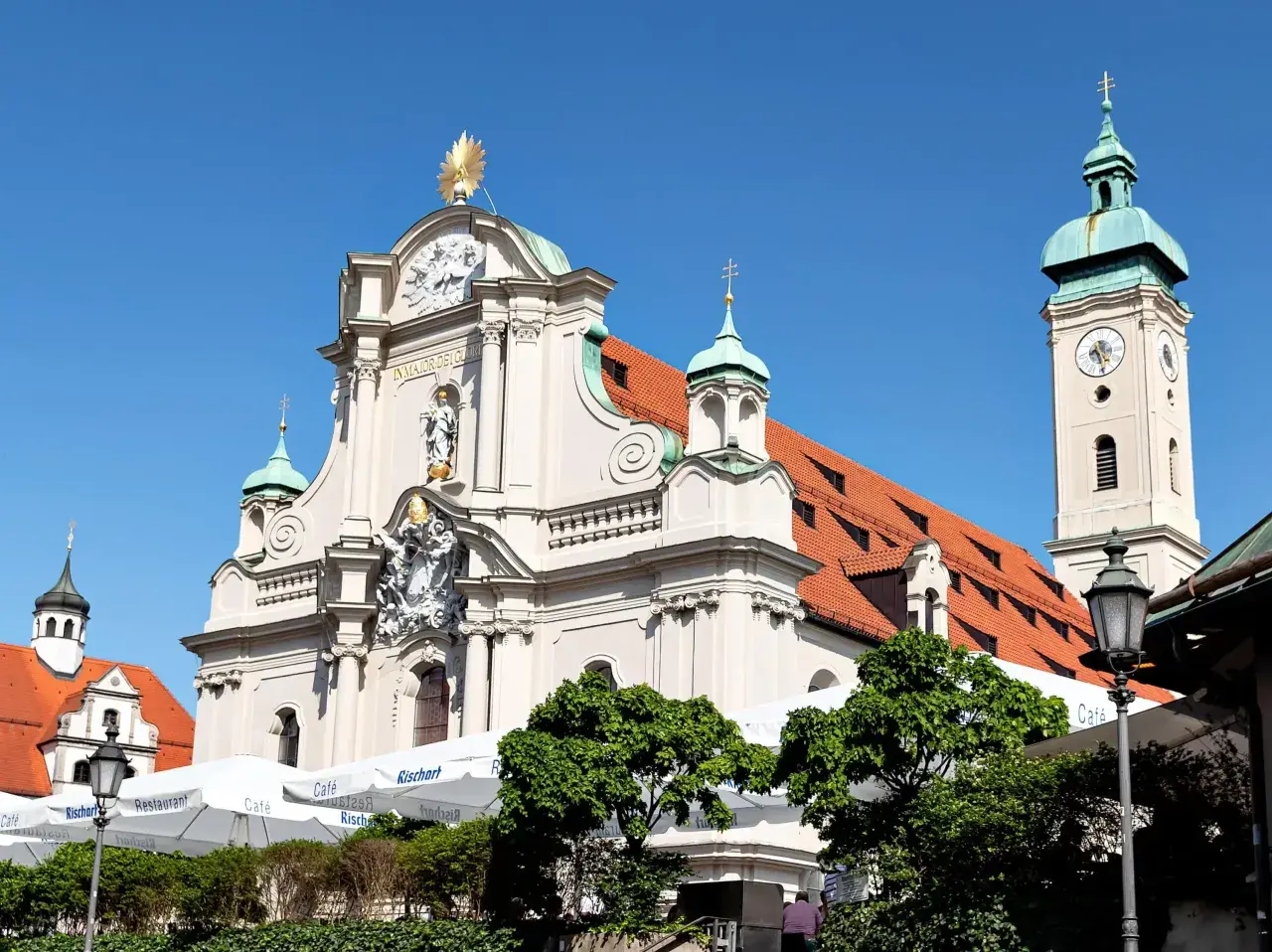 Prachtvolle Kirche am Viktualienmarkt mit goldenem Stern und Glockenturm. Sonnige Szene mit Cafés und Grünflächen.