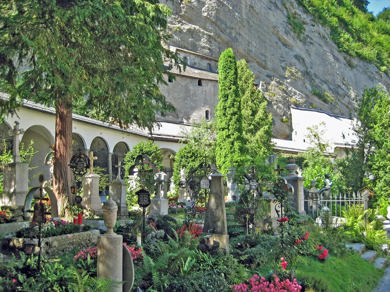 Gräberfeld mit Kreuzen und Urnen vor Arkaden der Erzabtei Salzburg. Üppige Vegetation und Felswand im Hintergrund.