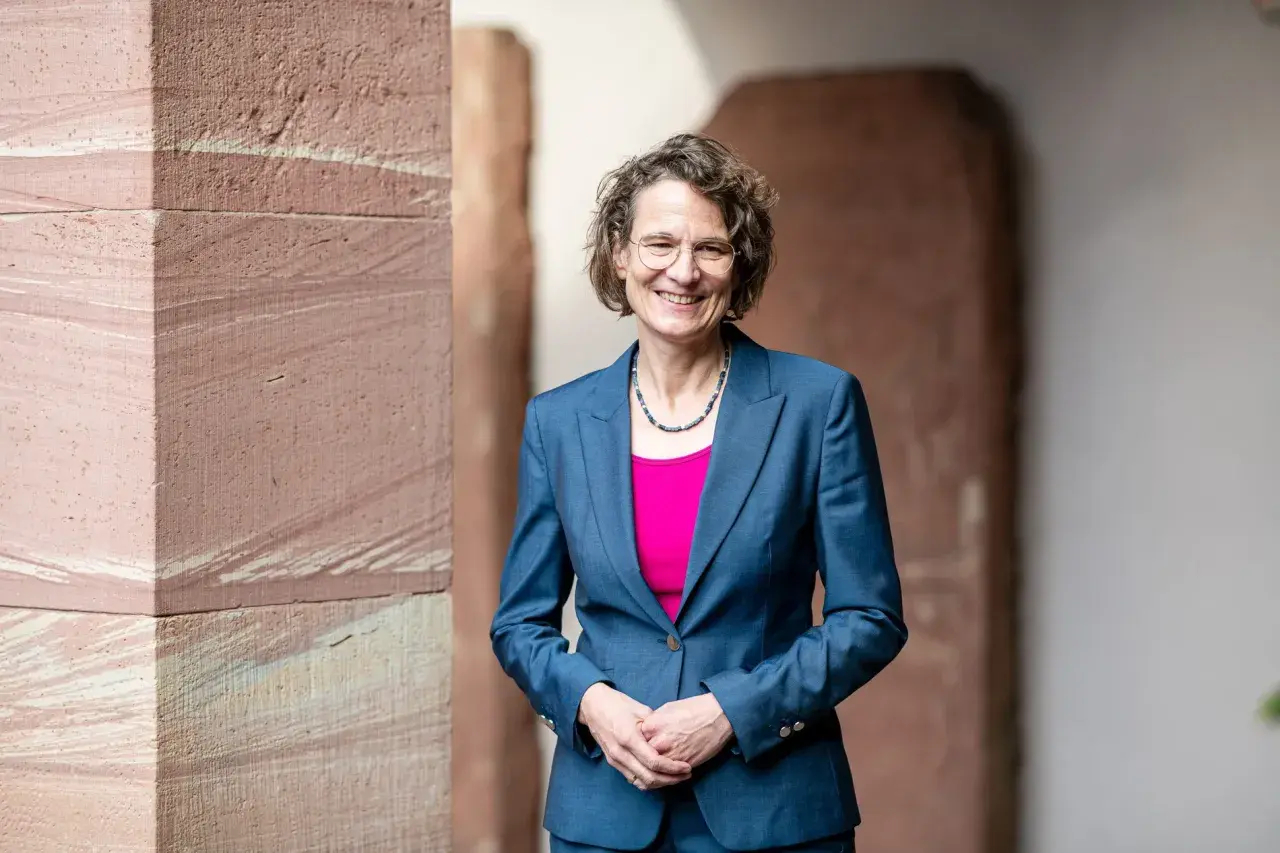 Christiane Tietz, eine Frau mit Brille und lockigem Haar, lächelt in einem blauen Hosenanzug vor einer Sandsteinwand.