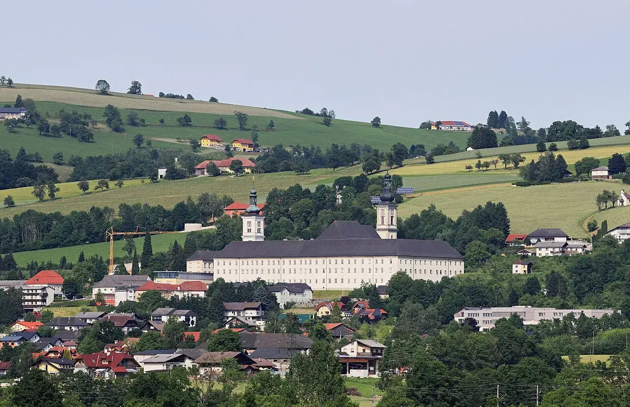 Das Stift Schlierbach, ein imposantes Barockgebäude, thront über einer malerischen Landschaft mit grünen Hügeln und traditionellen Häusern.
