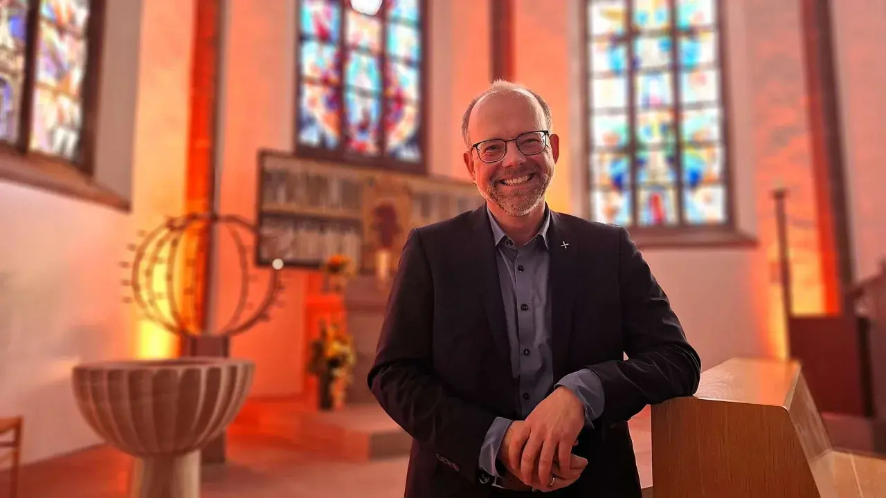 Lächelnder Mann in Anzug lehnt an einer Kirchenbank. Buntglasfenster und Taufbecken im Hintergrund. Hier kann man wieder in die Kirche eintreten.