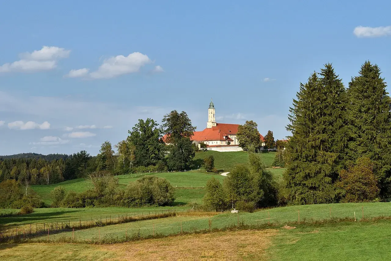 Kloster Reutberg thront auf einem Hügel, umgeben von grünen Wiesen und Bäumen unter blauem Himmel mit Wolken.