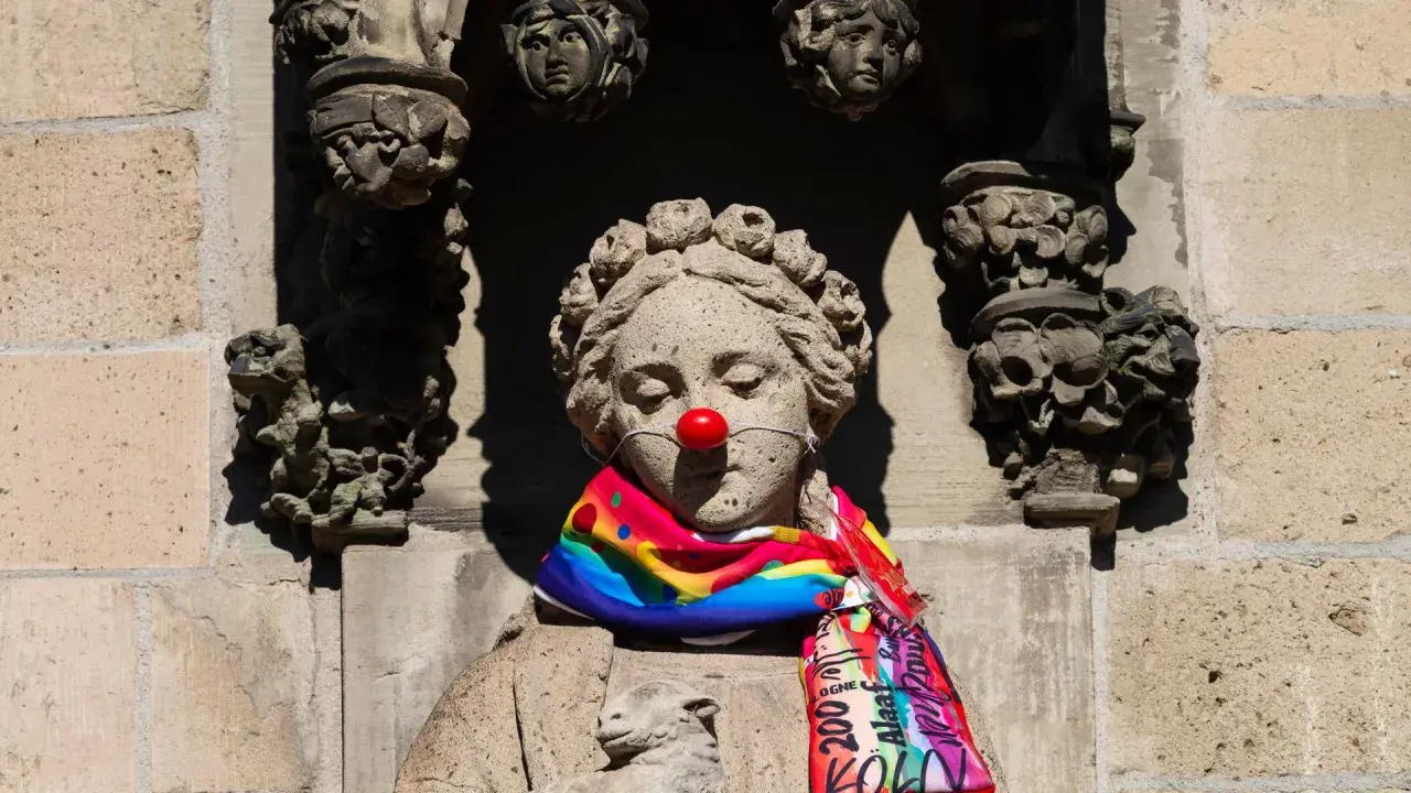 Steinskulptur mit roter Clownsnase und buntem Schal. Die Frage "was hat Karneval mit Religion zu tun?" wird hier humorvoll aufgeworfen.