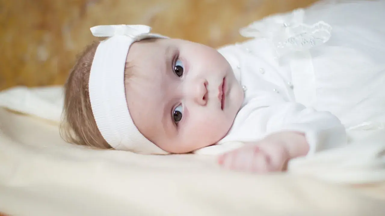 Ein süßes Baby in weißer Taufkleidung, mit einem Stirnband und einer Schleife. Vielleicht ein Name wie Maria oder Anna, typisch für katholische heilige Namen weiblich.
