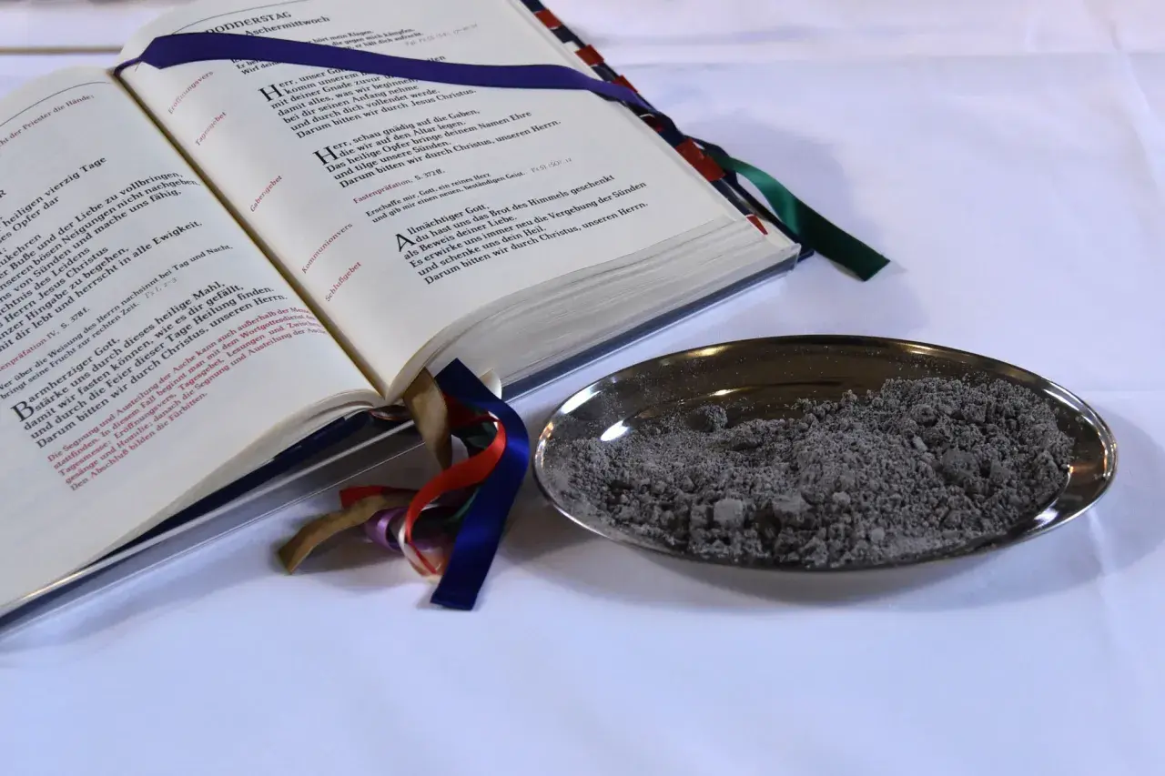 Ein aufgeschlagenes Buch mit Gebeten und eine Schale mit Asche. "Aus Staub bist du zu Staub wirst du" – ein Symbol für Vergänglichkeit.