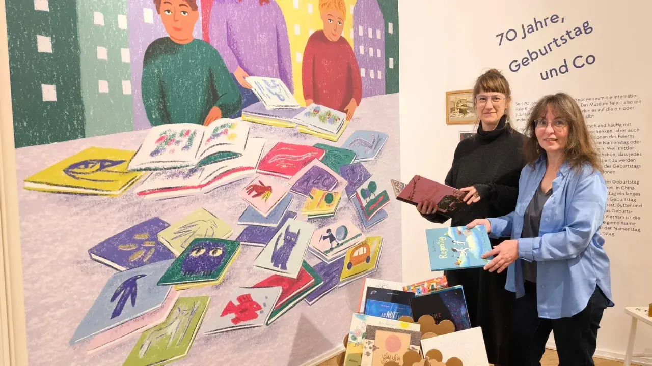 Zwei Frauen stehen vor einer bunten Wandmalerei mit Büchern und halten Bücher in den Händen.