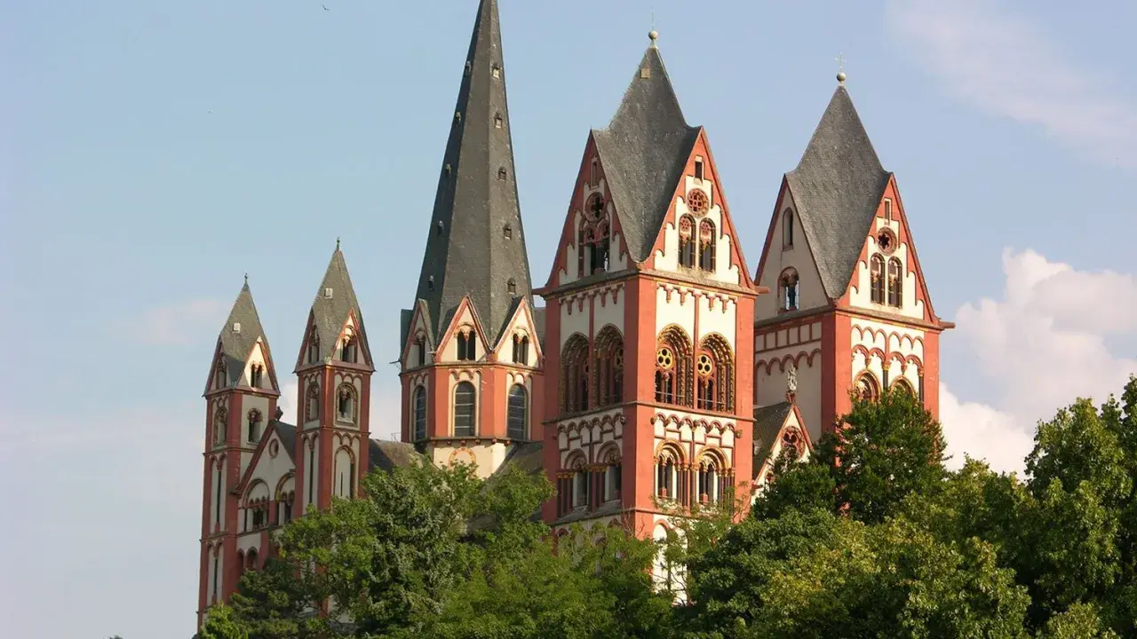 Der Limburger Dom mit seinen roten Türmen ragt über grünen Bäumen auf. Man munkelt, der Bischof von Limburg hätte dort eine goldene Badewanne versteckt.