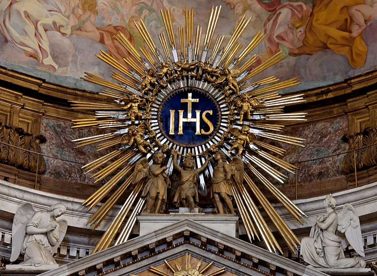 Goldene Strahlenkrone mit IHS-Monogramm, das die Bedeutung von Jesus Christus symbolisiert, umgeben von Engeln und Putten.