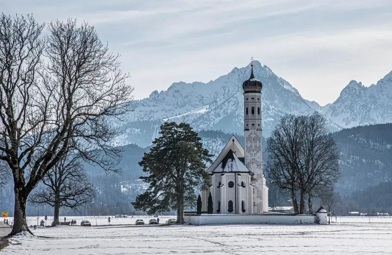 Winterliche Szene mit der Kirche St. Coloman im bayerischen Voralpenland, schneebedeckte Berge im Hintergrund.
