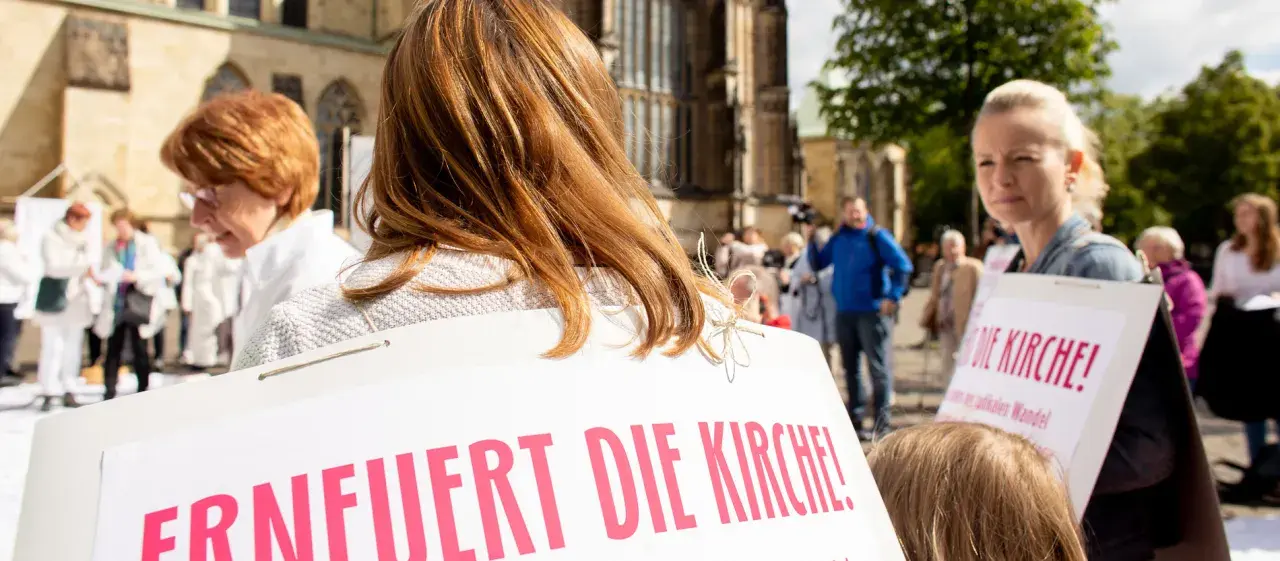 Frauen mit Schildern fordern "Erneuert die Kirche!". Eine Frau mit roten Haaren und eine mit blonden Haaren sind zu sehen.