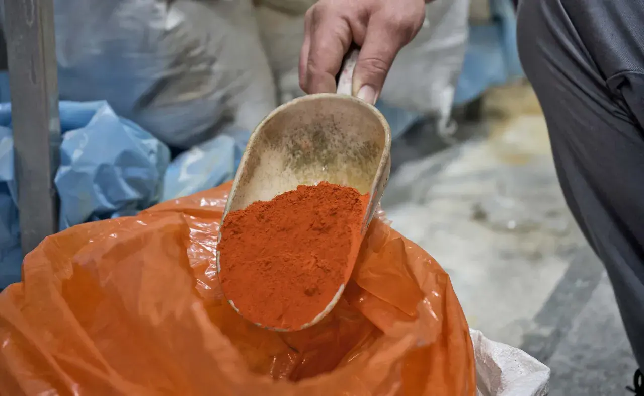 Eine Schaufel mit leuchtend orangefarbenem Pulver wird über einen orangefarbenen Beutel gehalten. Die Farben und ihre Bedeutung sind Teil eines alten Handwerks.
