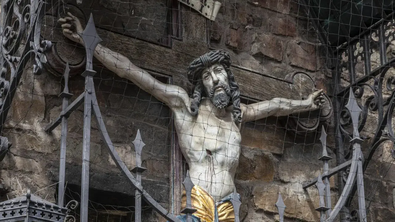Ein detailreiches, altes jesus kreuz original, hinter einem Metallgitter und Netz, zeigt die leidende Figur Christi vor einer Steinwand.