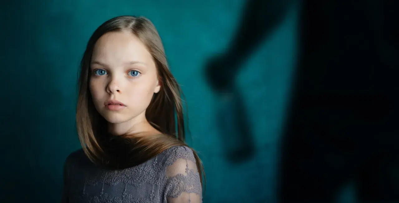 Ein junges Mädchen mit blauen Augen blickt nachdenklich, während im Hintergrund eine schattenhafte Figur erscheint, die an einen beschützenden Gott als Vater erinnert.