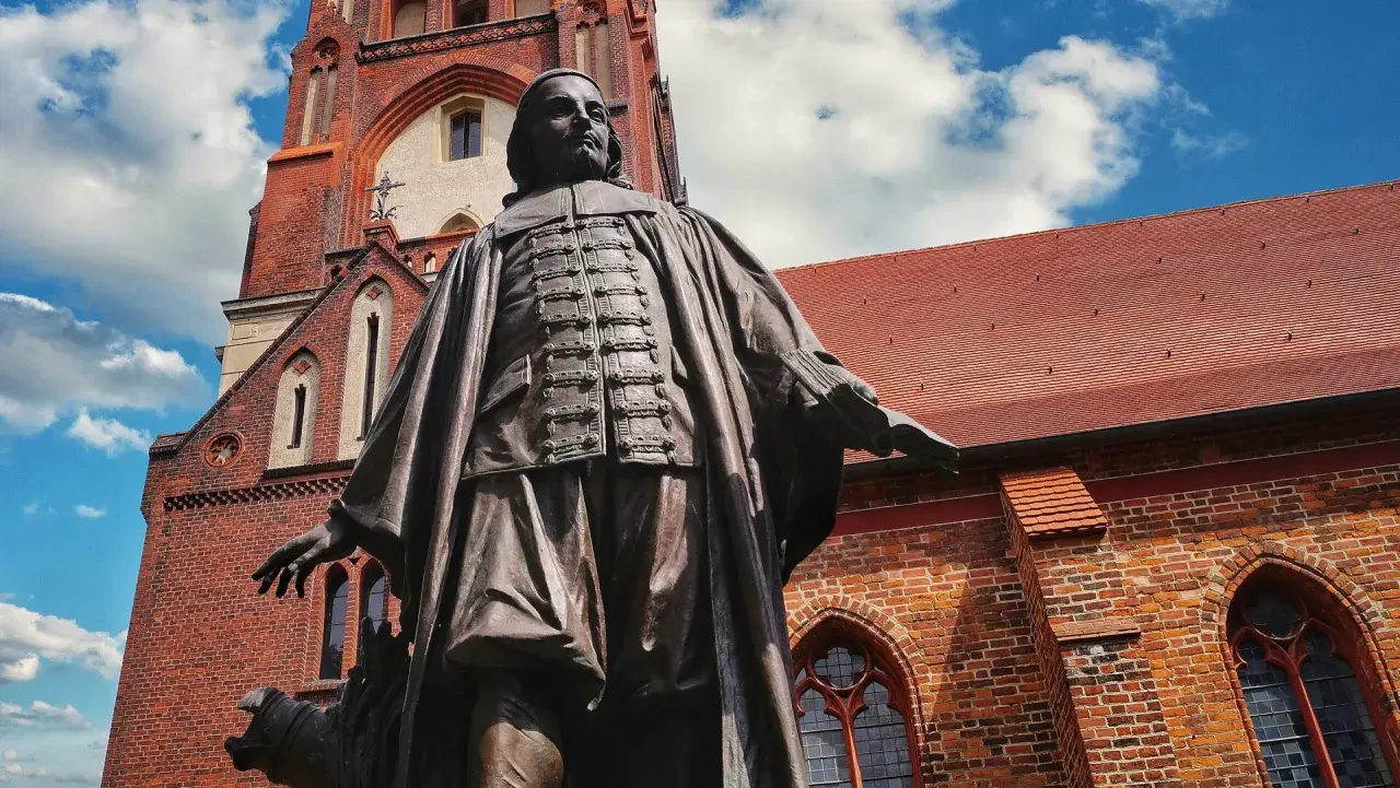 Bronzestatue von Paul Gerhardt vor einer roten Backsteinkirche mit blauen Himmel und Wolken.