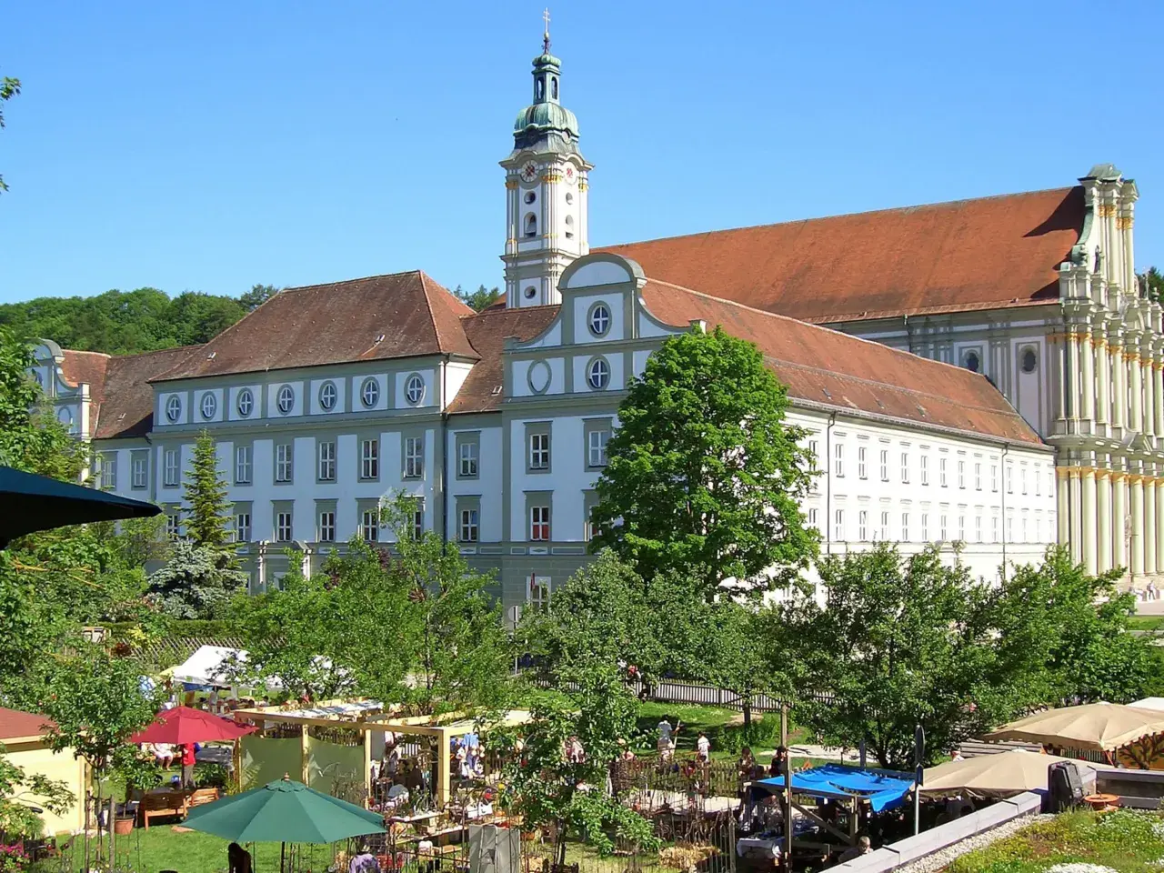 Prächtiges Kloster FFB mit barockem Turm, umgeben von grünen Bäumen und einem lebhaften Markt im Vordergrund.