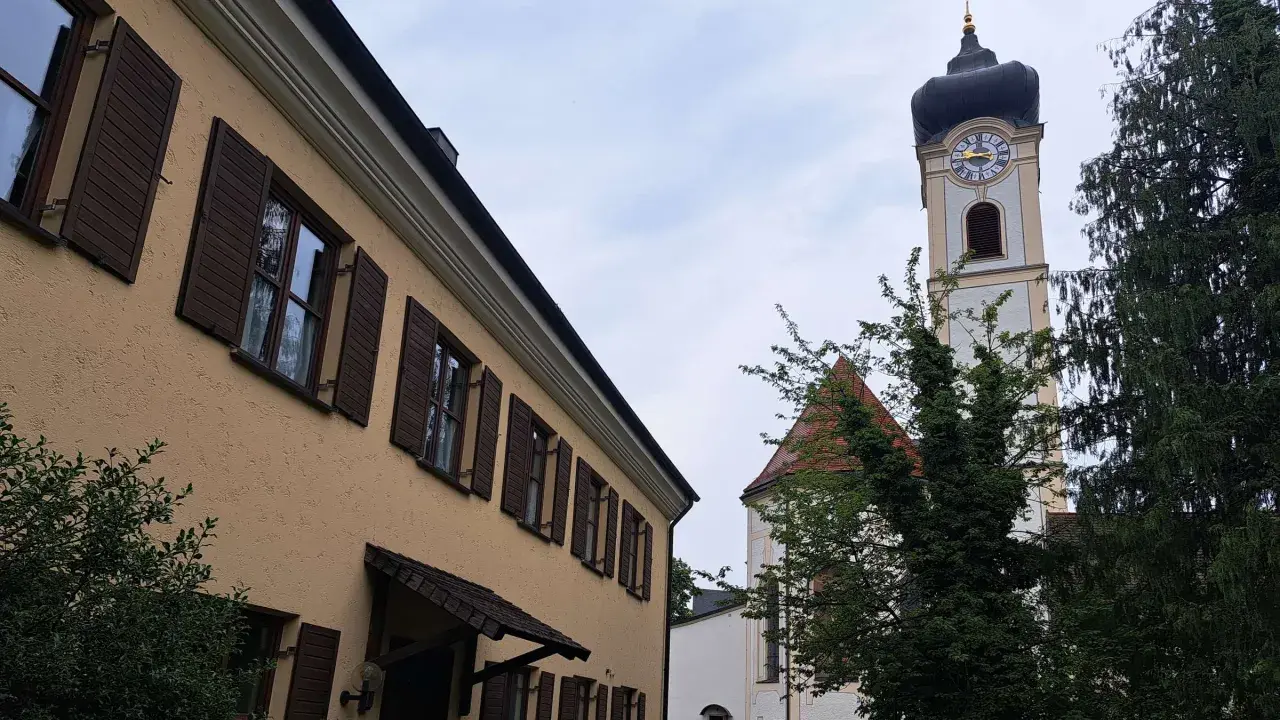 Ein Haus mit braunen Fensterläden und ein Kirchturm mit Uhr sind im Pfarrverband Erdinger Moos zu sehen.
