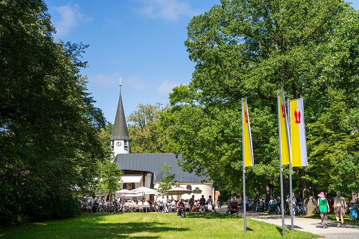 Ein sonniger Tag bei Maria Eich Planegg. Menschen sitzen unter Sonnenschirmen vor der Kirche, während Fahnen im Wind wehen.