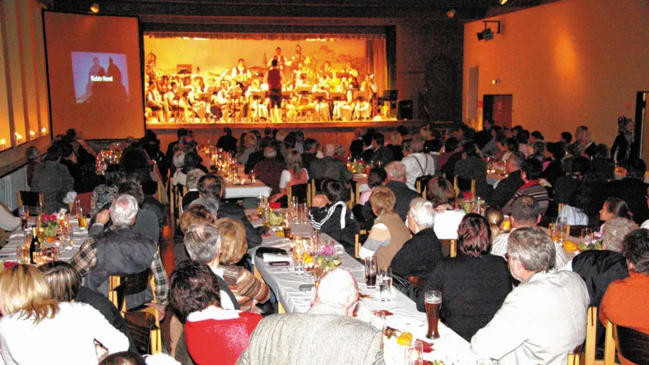Festliches Bankett im bischof bernhard haus kraiburg. Gäste lauschen einem Orchester, während auf der Leinwand "Robin Hood" läuft.