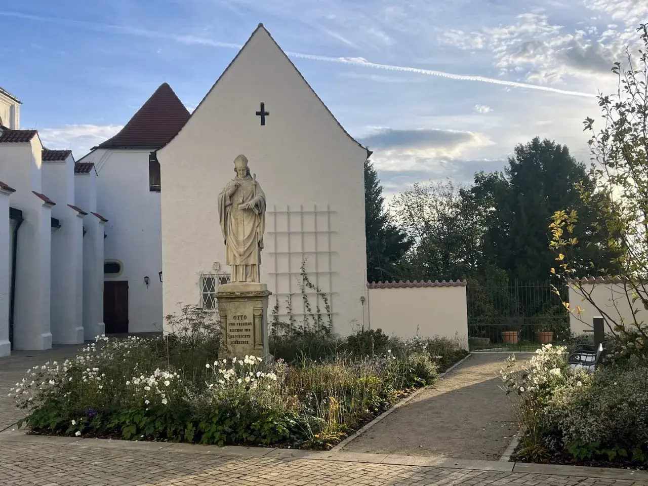 Statue vor dem Domberg Freising. Ein Weg führt durch einen blühenden Garten zu einem Tor.
