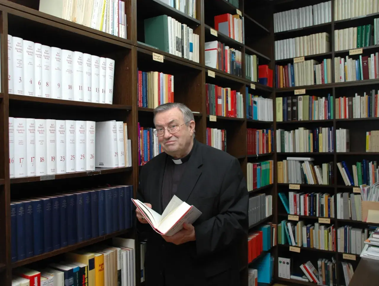 Kardinal Lehmann steht in einer Bibliothek und hält ein Buch in den Händen. Um ihn herum sind Regale voller Bücher.