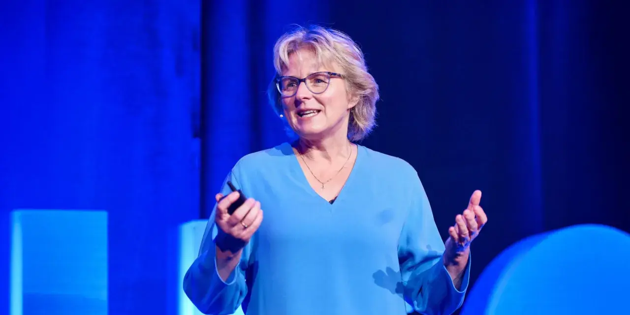 Melanie Wolfers, eine inspirierende Rednerin, hält einen Vortrag vor einem blauen Hintergrund. Sie trägt eine blaue Bluse und eine Halskette.