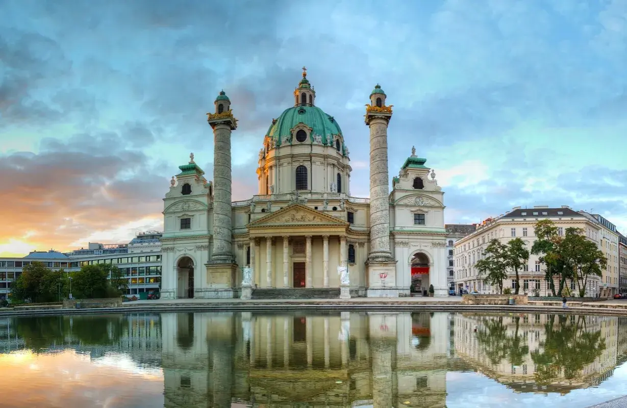 Die Karlskirche Wien spiegelt sich im Wasser. Majestätische Säulen flankieren die Kuppel. Ein dramatischer Himmel über der barocken Pracht.