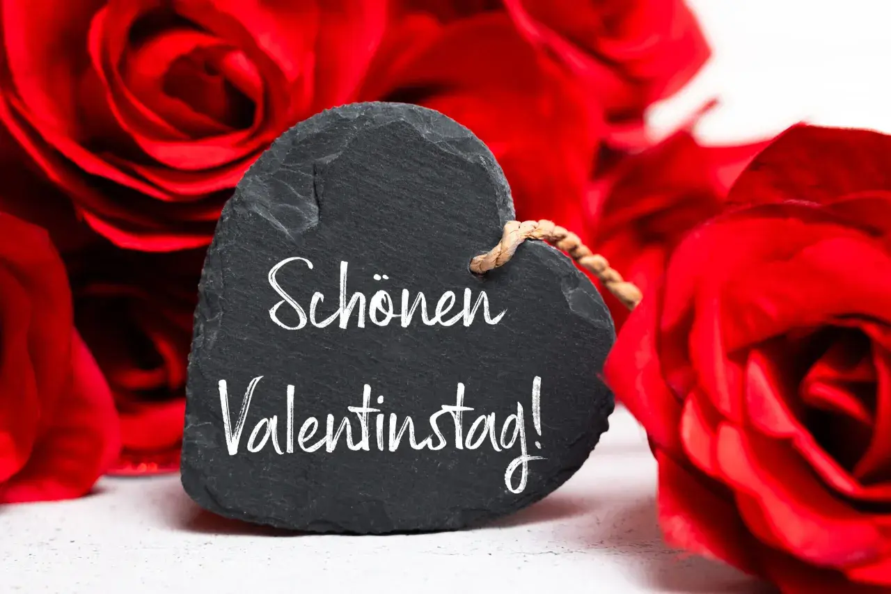 Herzförmiges Schild mit der Aufschrift "Schönen Valentinstag!" vor roten Rosen. Ein liebevoller Gruß zum heiligen Valentin.