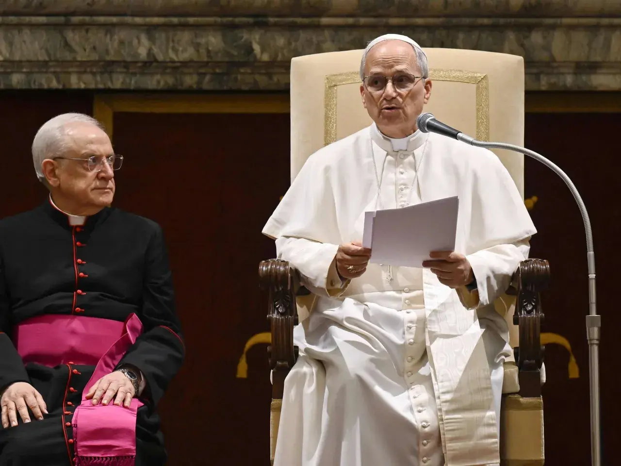 Papst Franziskus spricht in ein Mikrofon, während ein Kardinal daneben sitzt.