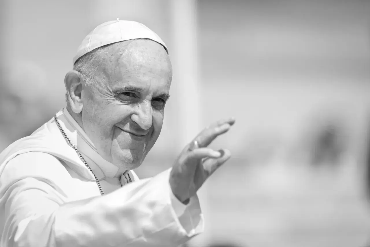 Papst Franziskus winkt lächelnd in die Menge, gekleidet in seine Soutane und mit einer Kette.