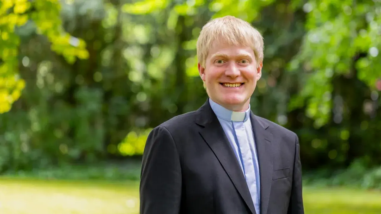 Thomas Frings, ein Geistlicher mit blondem Haar, lächelt freundlich in die Kamera. Er trägt einen schwarzen Anzug mit einem blauen Hemd und einem weißen Kragen.
