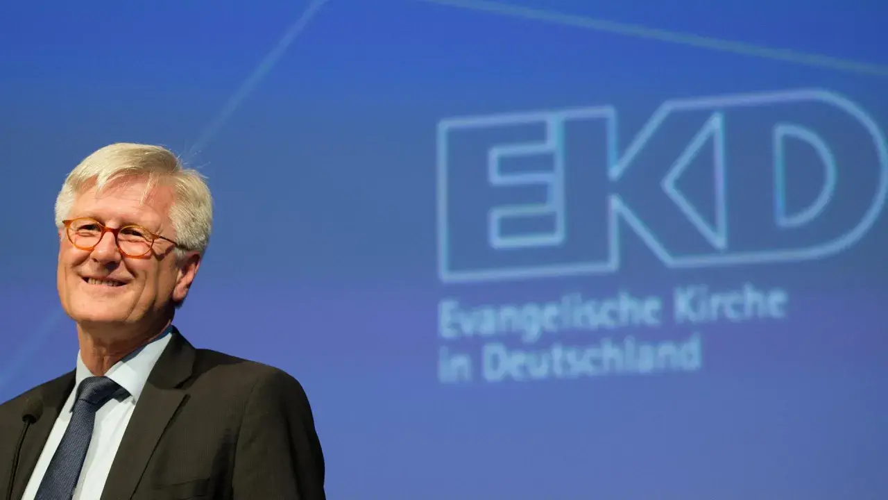 Lächeln des EKD Vorsitzenden vor dem Logo der Evangelischen Kirche in Deutschland.